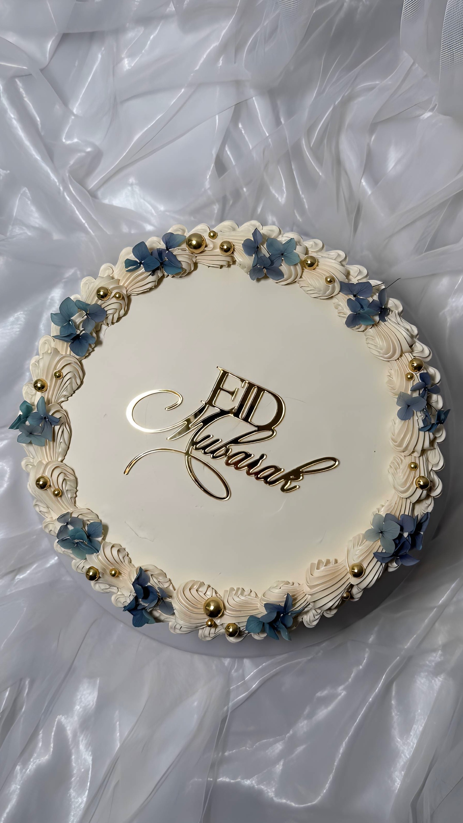 Round Eid cake with cream piping, blue flowers, and gold pearls